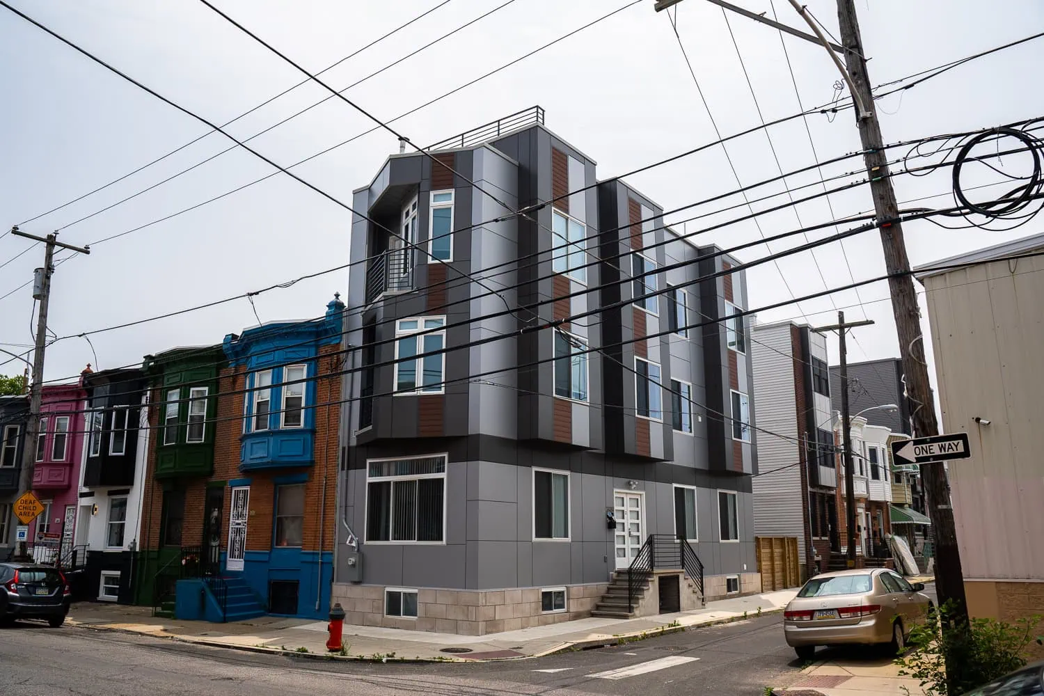 A modern three-story building with geometric design elements and uPVC windows stands on a street corner lined with older, colorful row houses. Power lines crisscross above, and various cars are parked nearby.